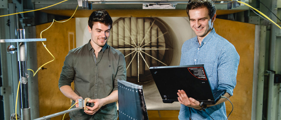 Two students are standing in the wind tunnel with a laptop